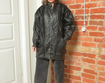 Könnte beinhalten: Eine schwarze Lederjacke mit Knopfleiste und Stehkragen. Die Jacke hat einen leicht oversized Schnitt und wird über einer dunkelgrauen Cordhose getragen.