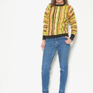 May include: A woman wearing a green, orange, and black striped sweater with a black crew neck and blue jeans. She is wearing black ankle boots.