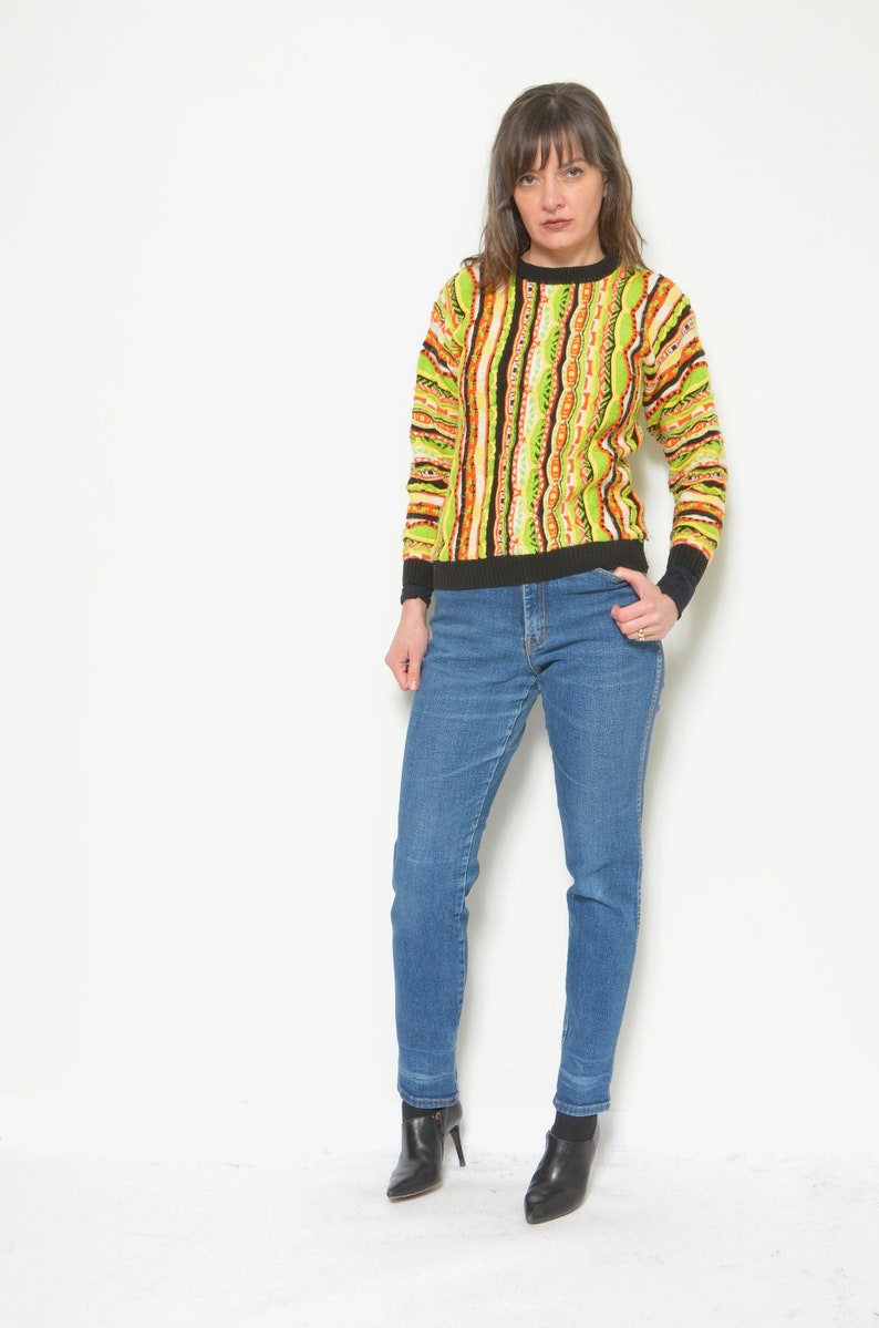 May include: A woman wearing a green, orange, and black striped sweater with a black crew neck and blue jeans. She is wearing black ankle boots.