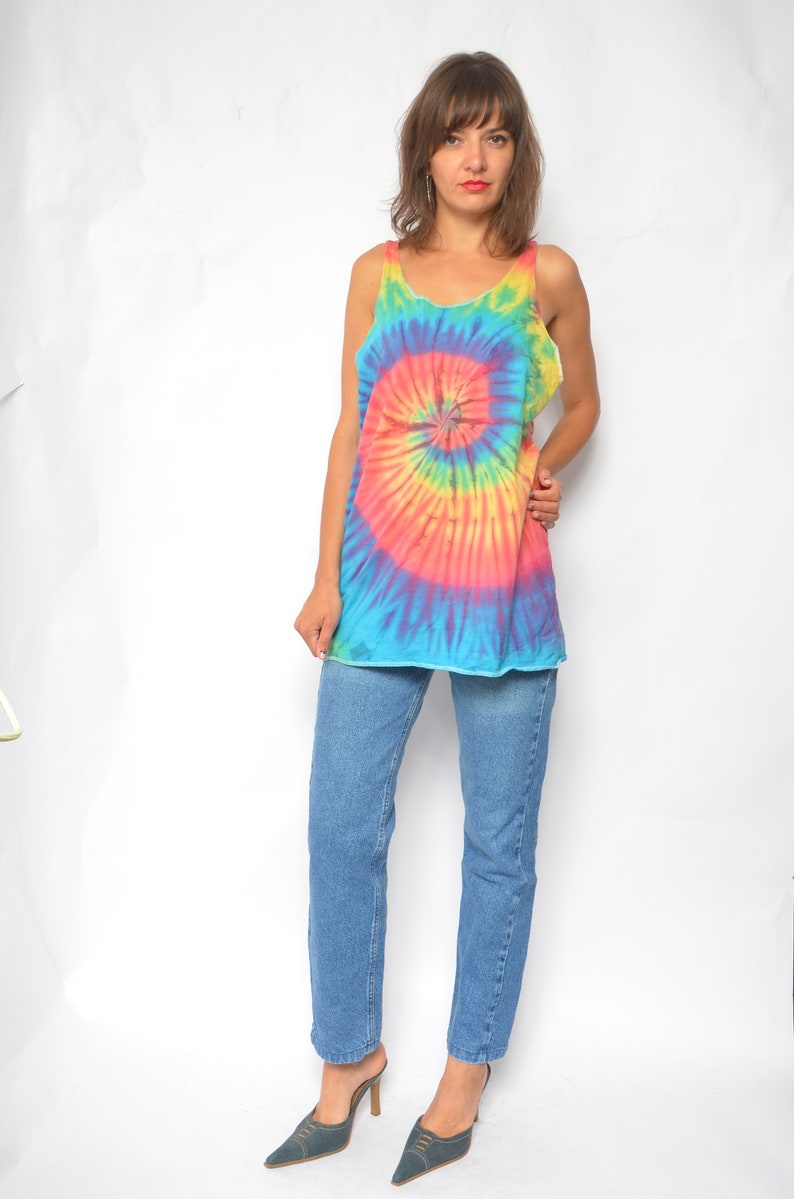 Puede incluir: Una mujer con un top sin mangas de tie-dye con un patr&oacute;n de espiral arco&iacute;ris y jeans azules. Lleva zapatos de tac&oacute;n azules.