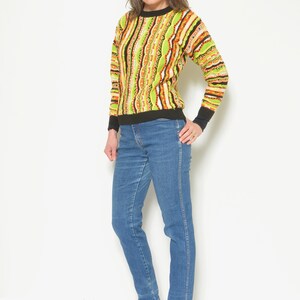 May include: A woman wearing a green, orange, and black striped sweater with blue jeans and black ankle boots.