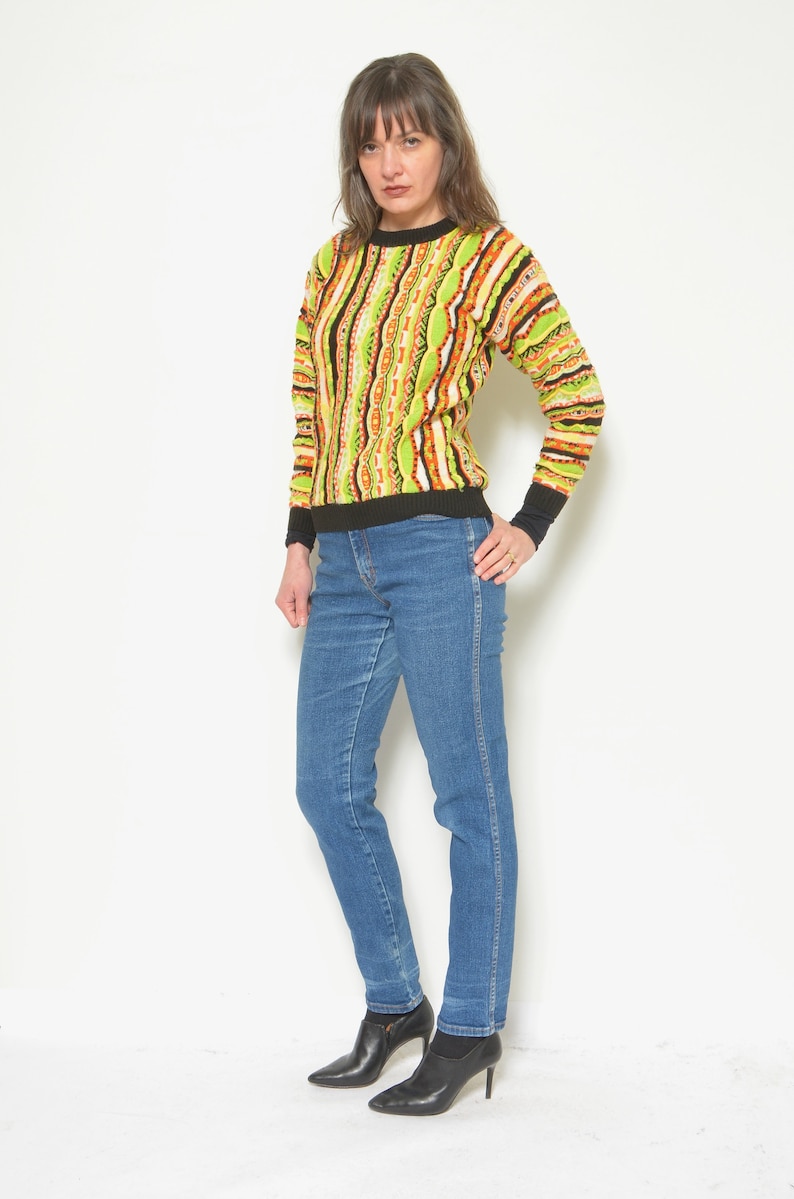 May include: A woman wearing a green, orange, and black striped sweater with blue jeans and black ankle boots.
