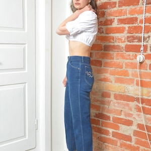 May include: A woman in a white crop top and high-waisted, dark blue jeans. The jeans feature decorative stitching on the back pocket. She is wearing patterned ankle boots with a pointed toe and a heel. The outfit is set against a brick wall.