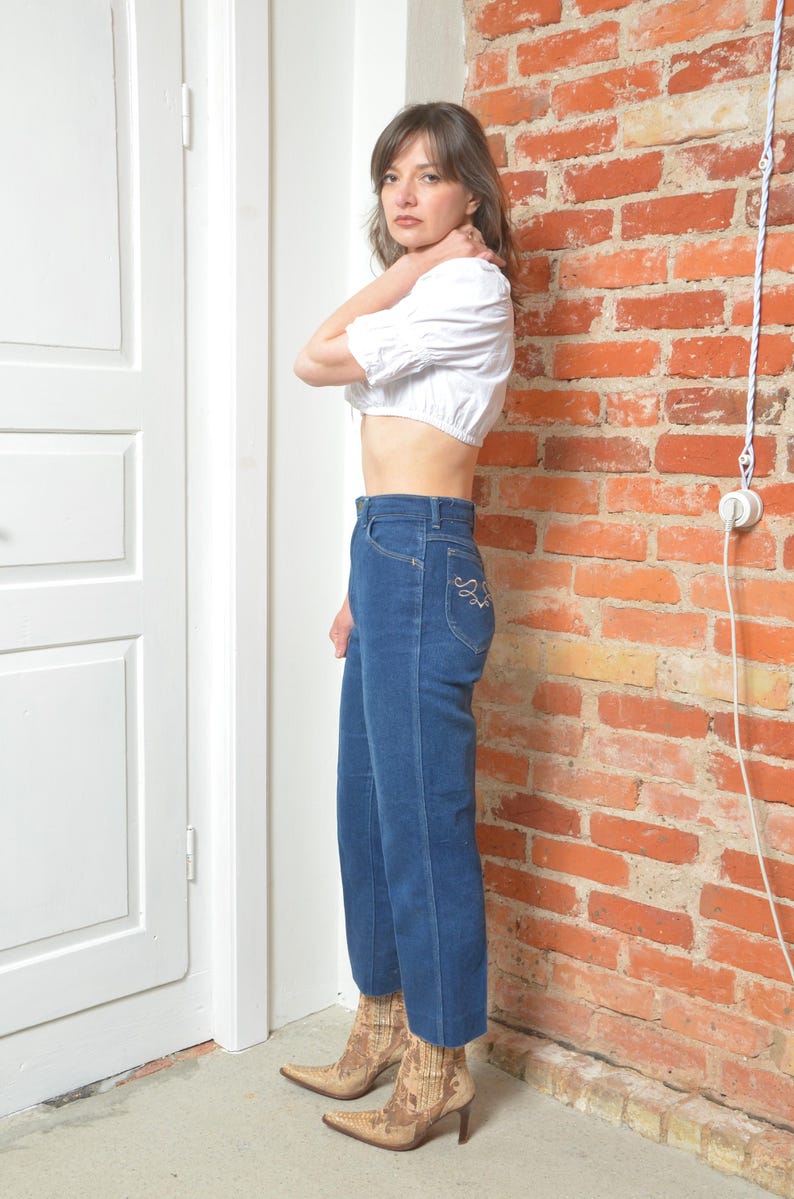 May include: A woman in a white crop top and high-waisted, dark blue jeans. The jeans feature decorative stitching on the back pocket. She is wearing patterned ankle boots with a pointed toe and a heel. The outfit is set against a brick wall.