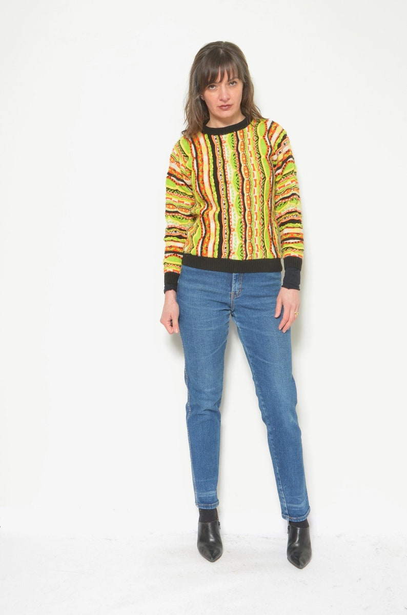 May include: A woman wearing a green, orange, and black striped sweater with a black crew neck and blue jeans.