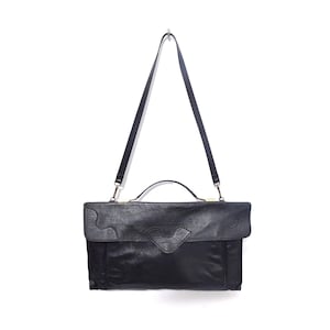 Könnte beinhalten: Schwarze Lederaktentasche mit Klappenverschluss und oberem Griff. Die Klappe hat ein dekoratives Wellendesign. Die Tasche hat einen langen, verstellbaren Schultergurt. Die Aktentasche hängt vor einem weißen Hintergrund.