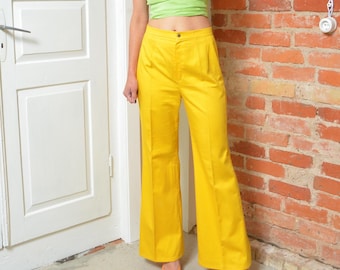 Puede incluir: Pantalones amarillos brillantes de pierna ancha con cintura alta. Los pantalones se combinan con un top verde claro y zapatos naranjas. Los pantalones tienen un diseño clásico y están hechos de una tela suave. El conjunto se encuentra frente a una puerta blanca y una pared de ladrillo rojo.