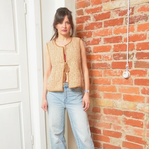 May include: A tan and cream-colored vest with a textured pattern. The vest has a brown trim and a tie closure. The person is wearing light blue jeans. The background is a red brick wall and a white door.