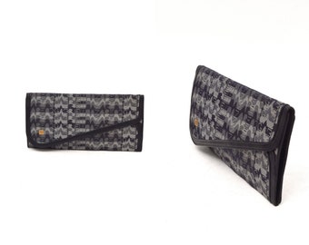 May include: A black and gray patterned clutch purse with a flap closure. The purse has a small, rectangular, gold-colored accent on the front.