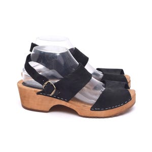 Pode incluir: Uma caneca de cerâmica branca com uma alça preta e o texto "But First, Coffee" em negrito preto. A caneca está sobre um fundo desfocado, sugerindo um ambiente acolhedor.