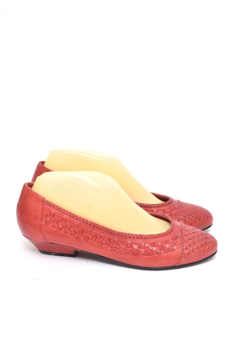 May include: A pair of coral-colored flats with a woven design on the toe and vamp. The shoes have a low heel and a rounded toe. The shoes are displayed on a white background.