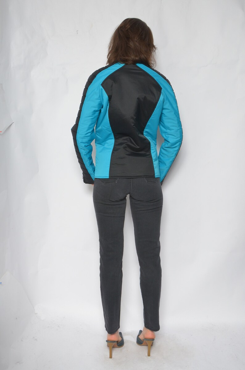 May include: A black and turquoise jacket with a zipper closure. The jacket has a quilted design and is worn over a pair of dark denim jeans.