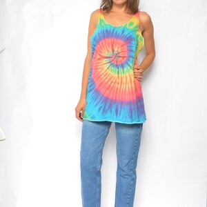 Puede incluir: Una mujer con un top sin mangas de tie-dye con un patr&oacute;n de espiral arco&iacute;ris y pantalones vaqueros azules. Lleva zapatos de tac&oacute;n azul oscuro.