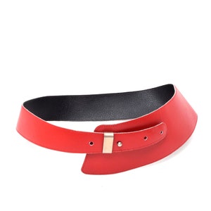 Op de afbeelding: Een rode leren riem met een gouden gesp. De riem is ontworpen met een unieke asymmetrische vorm.