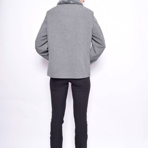 Op de afbeelding: Een grijze blazer met een nep bontkraag. De blazer is open en de persoon draagt een zwarte broek en zwarte schoenen.