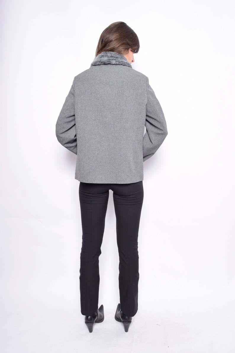 Op de afbeelding: Een grijze blazer met een nep bontkraag. De blazer is open en de persoon draagt een zwarte broek en zwarte schoenen.