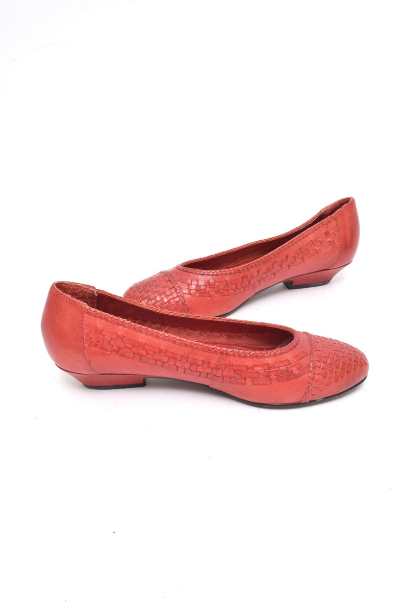 May include: A pair of coral-colored flats with a woven design on the toe and vamp. The shoes have a low heel and a rounded toe. The shoes are made of leather.