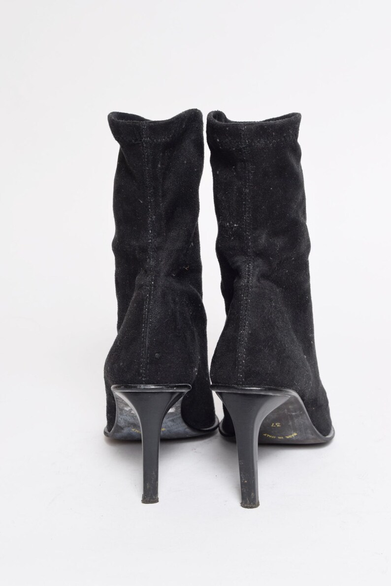 May include: A pair of black suede heeled boots with a pointed toe. The boots have a stretchy upper and a high heel.