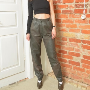 Könnte beinhalten: Olivgrüne Lederhose mit hoher Taille und maßgeschneiderter Passform. Die Hose hat einen Knopfverschluss und verfügt über Paneeldetails an den Beinen. Das Model trägt ein schwarzes One-Shoulder-Top und braune Schuhe.