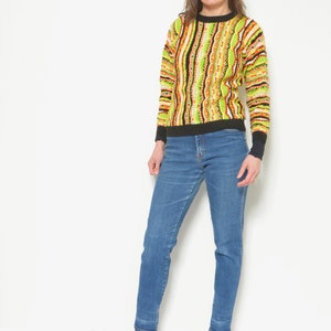 May include: A woman wearing a green, orange, and black striped sweater with blue jeans and black heeled ankle boots.