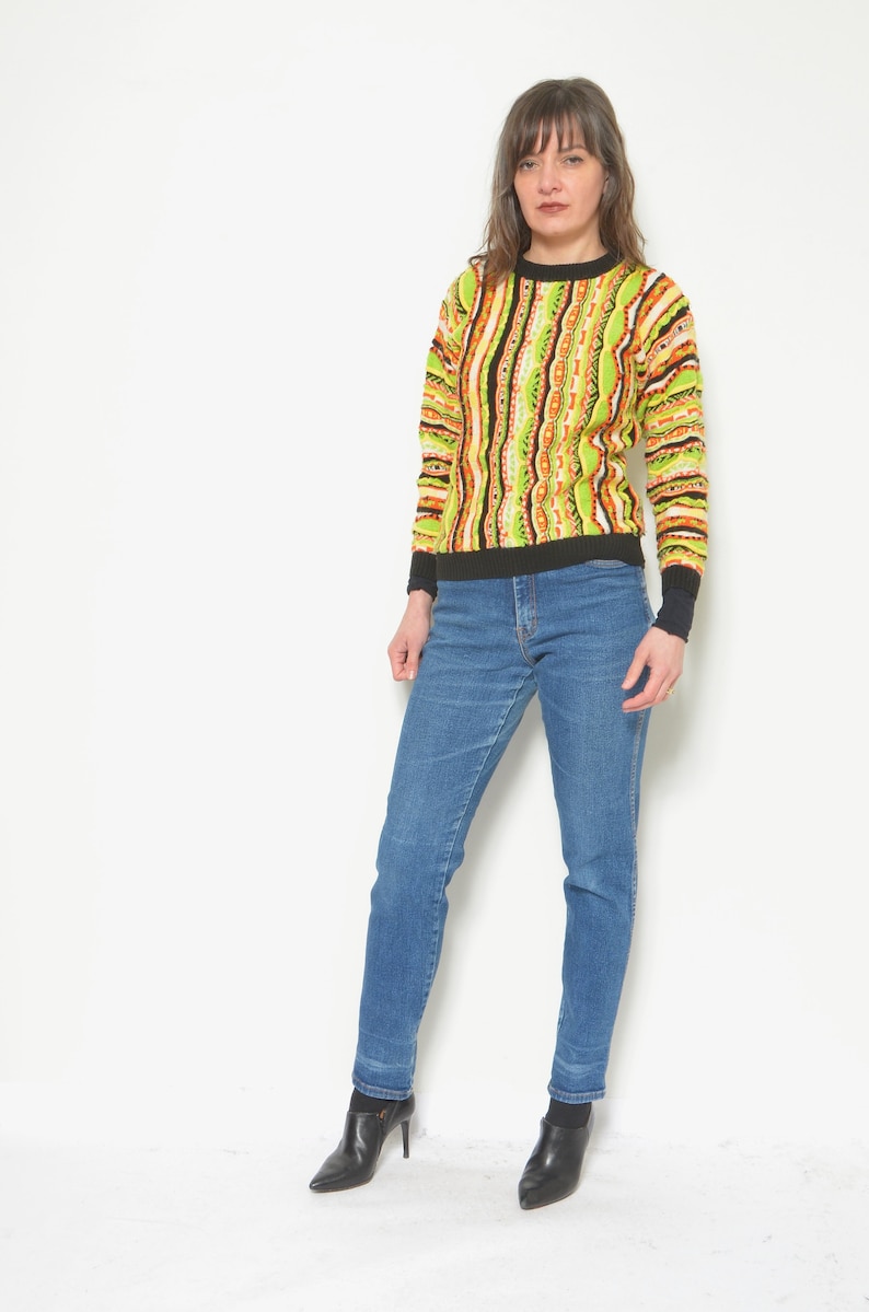 May include: A woman wearing a green, orange, and black striped sweater with blue jeans and black heeled ankle boots.
