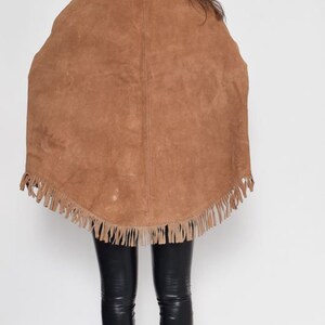 May include: A brown suede cape with fringe trim. The cape is worn over black leggings and black heeled boots.