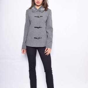 Op de afbeelding: Een grijze blazer met een nep bontkraag en vier zwarte gespen. De blazer heeft lange mouwen en een aansluitende pasvorm. Het model draagt een zwarte broek en zwarte laarzen.