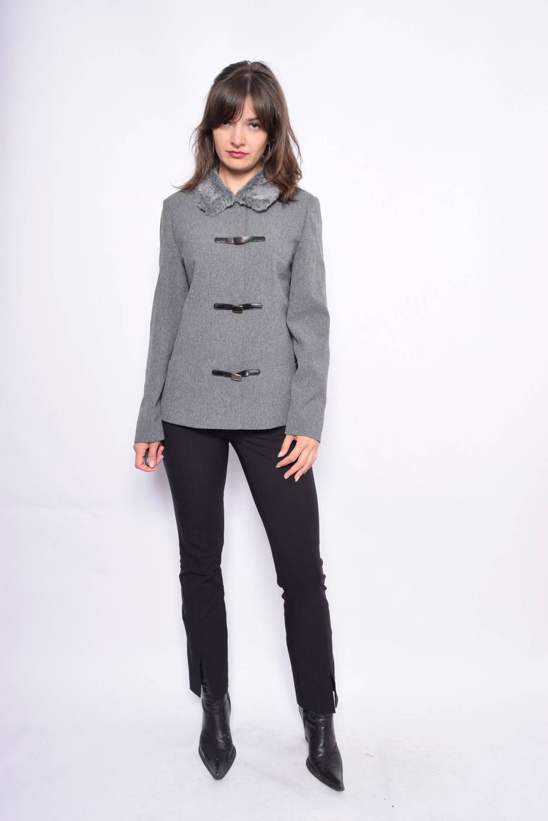 Op de afbeelding: Een grijze blazer met een nep bontkraag en vier zwarte gespen. De blazer heeft lange mouwen en een aansluitende pasvorm. Het model draagt een zwarte broek en zwarte laarzen.