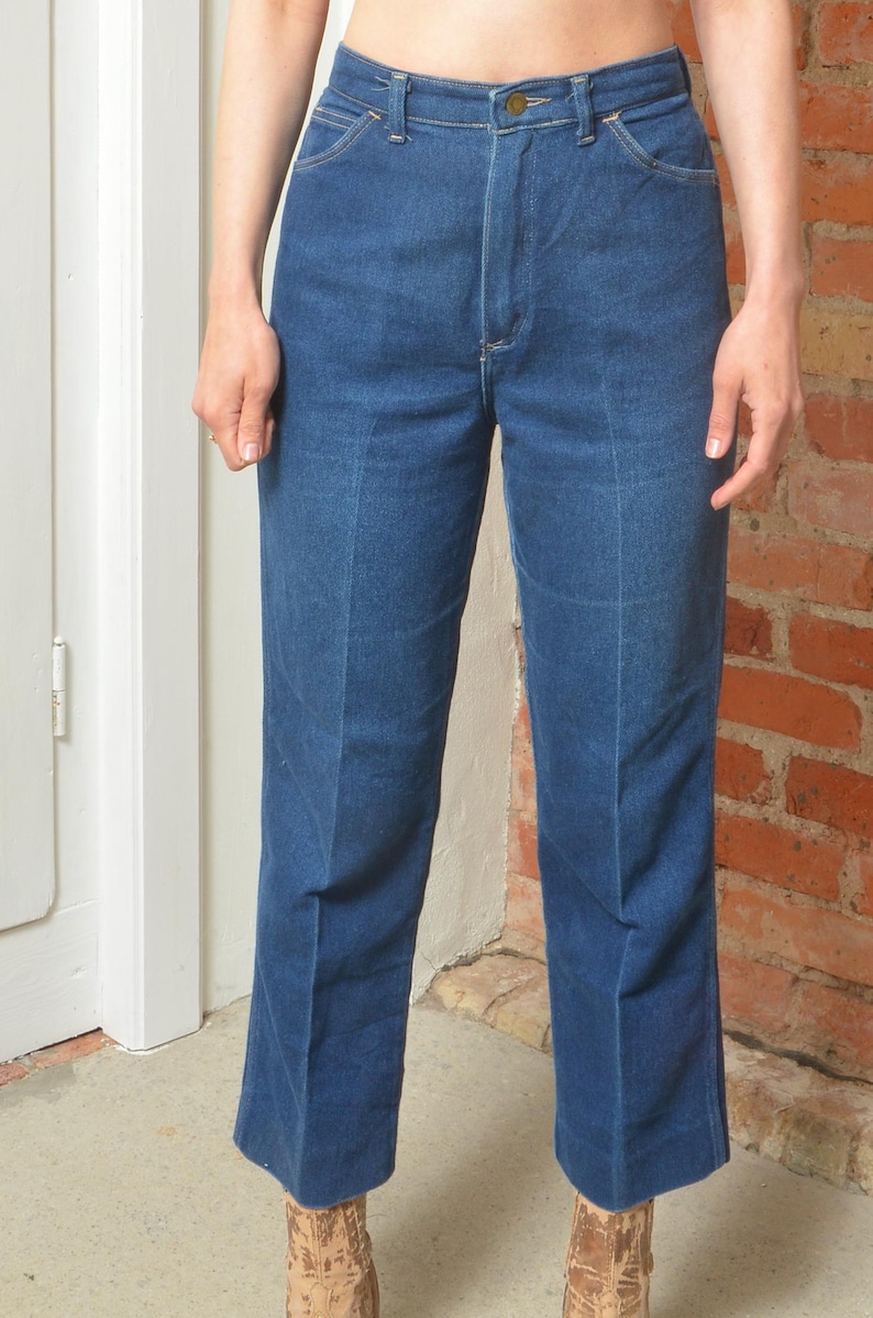 May include: Dark blue denim jeans with a high waist and a straight leg. The jeans feature a button closure and pockets. The model is wearing the jeans. The background is a brick wall and a white door.