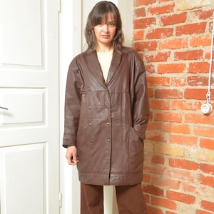 Könnte beinhalten: Ein brauner Ledermantel mit Reverskragen, Knopfverschlüssen und Taschen. Der Mantel ist knielang und hat ein klassisches, maßgeschneidertes Design. Er wird über einem cremefarbenen Oberteil und einer braunen Hose getragen.