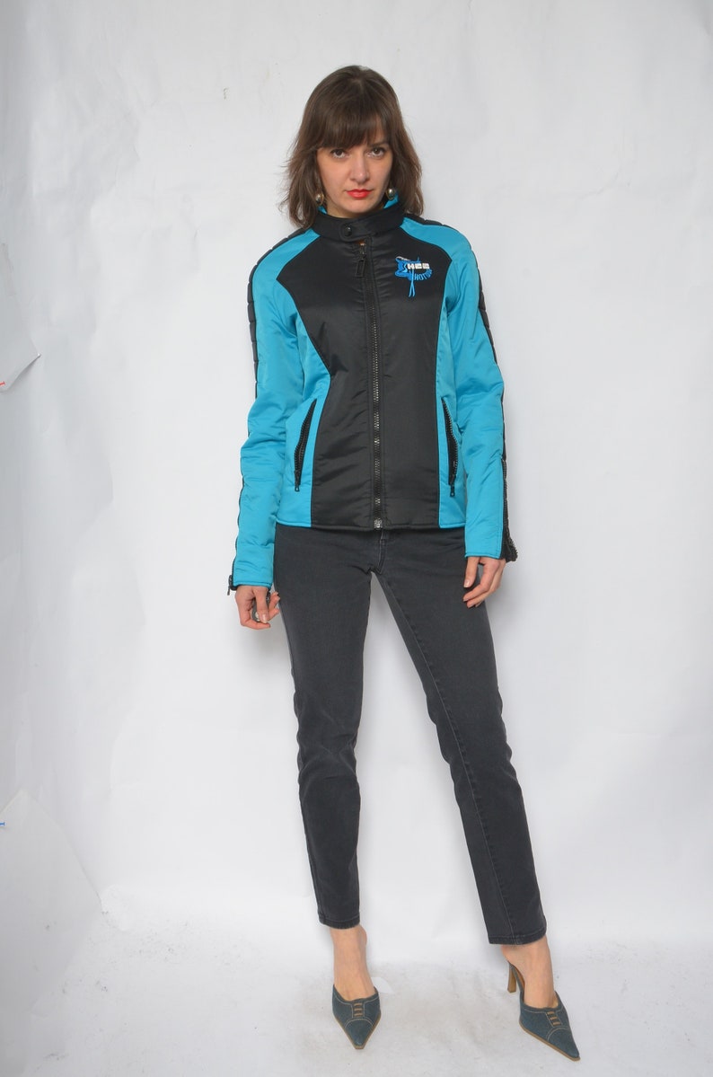 May include: A black and blue jacket with a zipper closure and a logo that says "HBS" on the front. The jacket has a stand-up collar and long sleeves. The person wearing the jacket is also wearing black jeans and blue shoes.