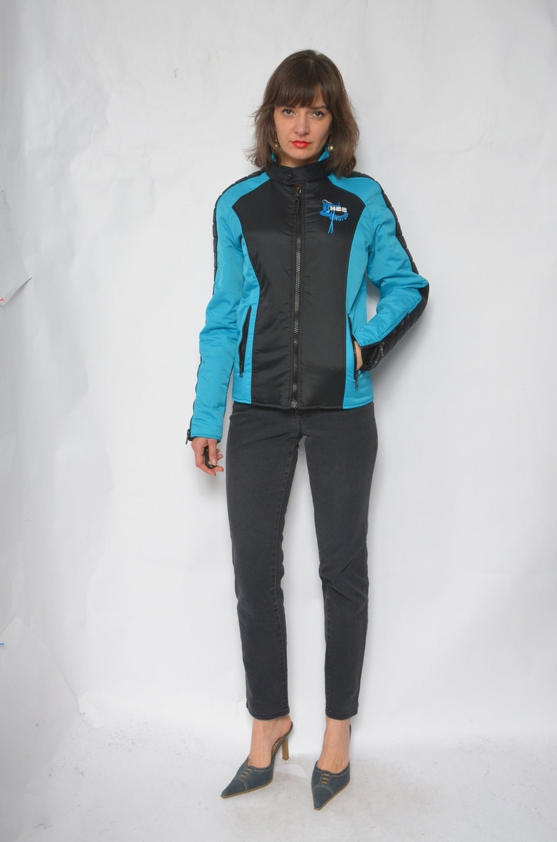 May include: A woman wearing a black and blue jacket with a zipper closure. The jacket has a blue and black color block design. She is wearing black jeans and blue heeled shoes.