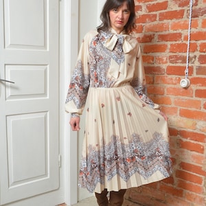 Può includere: Abito vintage color crema con gonna a pieghe e maniche lunghe. L'abito presenta un motivo floreale e geometrico nei toni del marrone, grigio e arancione. Ha un fiocco allo scollo e una vita aderente, creando una silhouette classica.