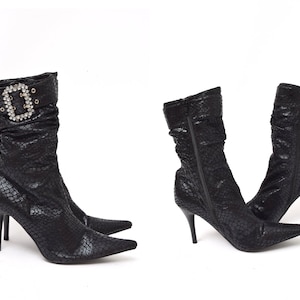 Könnte beinhalten: Ein Paar schwarze Stiefeletten mit Absatz und spitzer Zehenpartie. Ein Stiefel hat eine dekorative Schnalle mit Strasssteinen. Die Stiefeletten haben ein Schlangenledermuster.