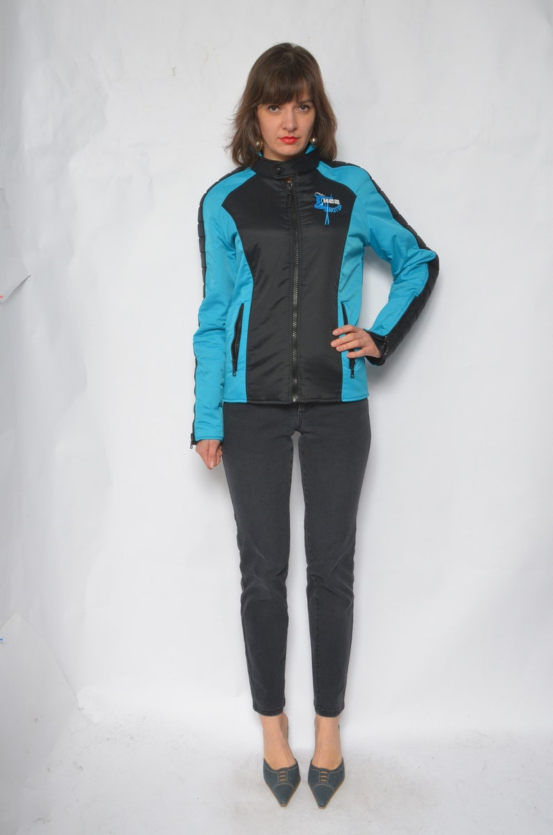 May include: A woman wearing a black and blue jacket with a zipper closure and a logo on the chest. She is wearing black pants and blue shoes.
