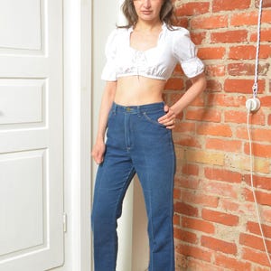 May include: A woman in a white crop top and high-waisted, straight-leg blue jeans. She is wearing patterned ankle boots with a pointed toe and a heel. The jeans are a classic style. The woman is standing near a brick wall.