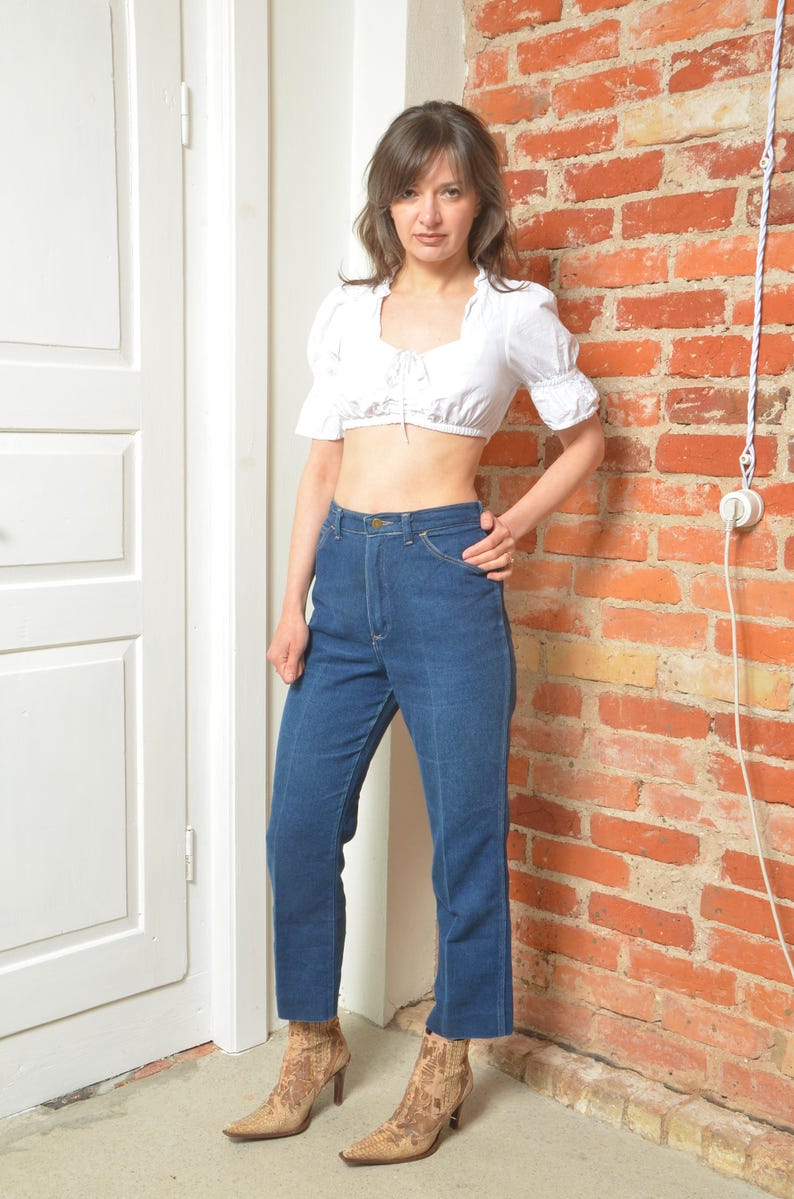 May include: A woman in a white crop top and high-waisted, straight-leg blue jeans. She is wearing patterned ankle boots with a pointed toe and a heel. The jeans are a classic style. The woman is standing near a brick wall.