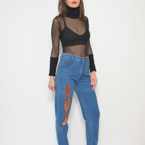 May include: Blue denim jeans with an embroidered design in shades of brown. The jeans have a high waist and a straight leg. The model is wearing a black turtleneck top and black boots.