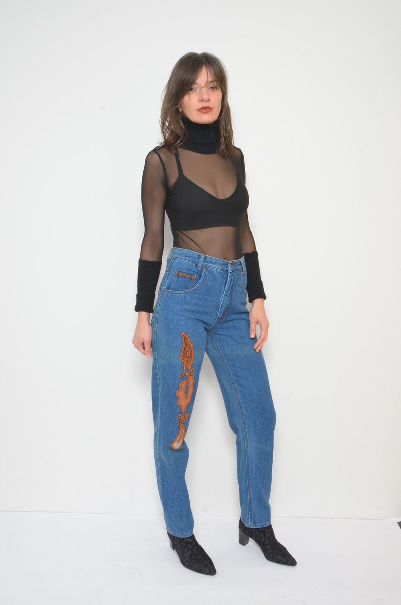 May include: Blue denim jeans with an embroidered design in shades of brown. The jeans have a high waist and a straight leg. The model is wearing a black turtleneck top and black boots.