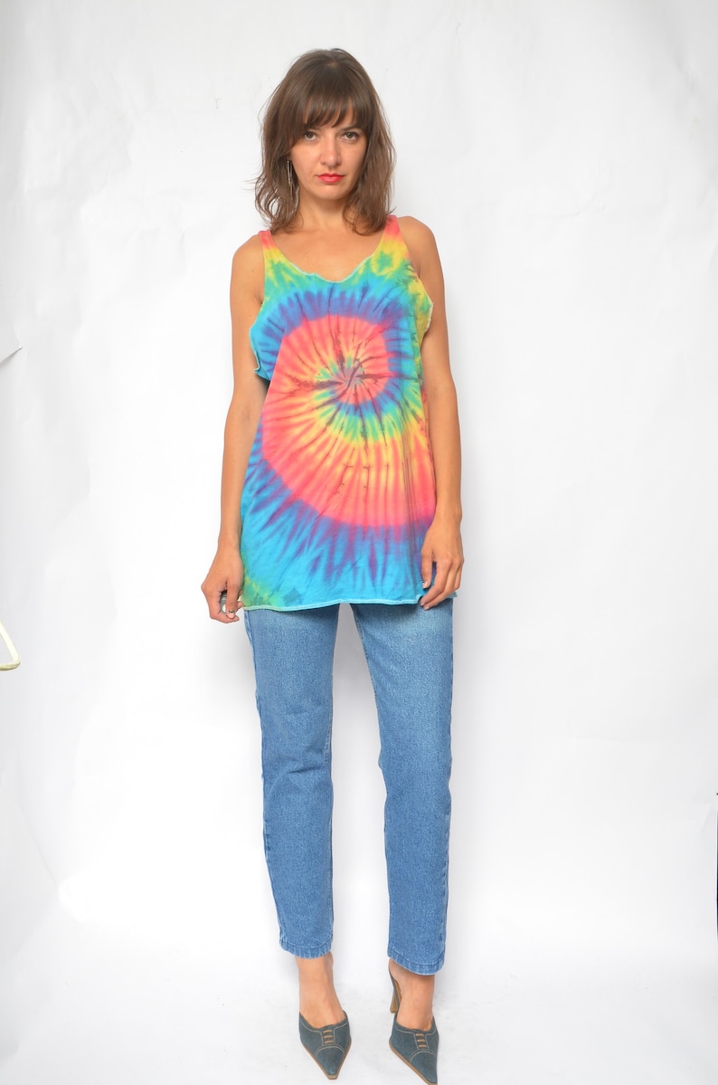 Puede incluir: Un top sin mangas de tie-dye con un patr&oacute;n de espiral arco&iacute;ris. El top es sin mangas y tiene un escote redondo. La modelo lleva pantalones vaqueros azules y tacones azul oscuro.