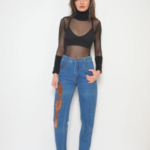 May include: A woman wearing a black turtleneck top and blue jeans. The jeans have an embroidered design on the front. The top is sheer, and the woman is also wearing a black bra. She is wearing black boots.