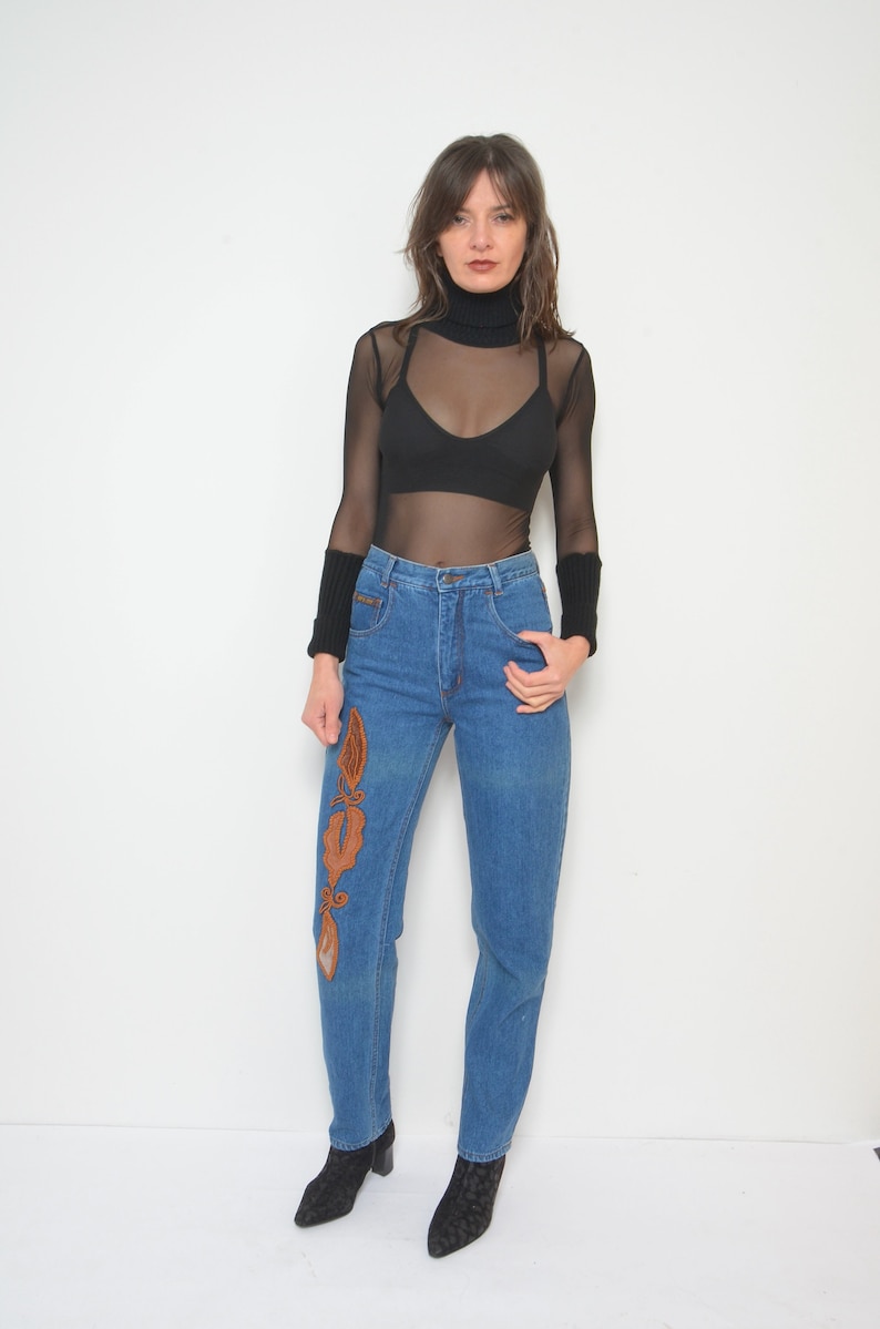 May include: A woman wearing a black turtleneck top and blue jeans. The jeans have an embroidered design on the front. The top is sheer, and the woman is also wearing a black bra. She is wearing black boots.