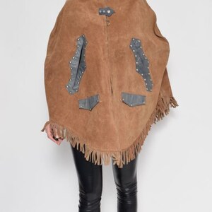 May include: A brown suede poncho with fringe and black leather accents. The poncho has a zipper closure and a stand-up collar.
