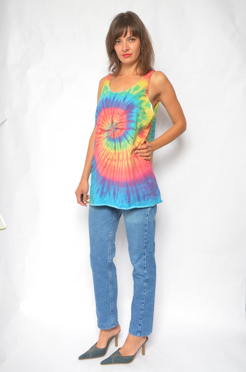 Puede incluir: Una mujer con un top sin mangas de tie-dye con un dise&ntilde;o de espiral azul, rosa, amarillo y naranja. Tambi&eacute;n lleva pantalones vaqueros azules y zapatos de tac&oacute;n azul oscuro.
