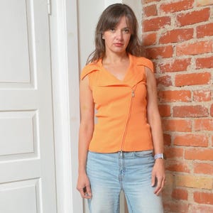 May include: An orange sleeveless top with a diagonal zipper closure. The top has a V-neck and a ribbed texture. The person is wearing light blue jeans. The background includes a brick wall and a white door.