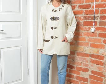 May include: Cream-colored long-sleeve coat with dark brown toggle closures and pockets. The coat is paired with light blue jeans and orange heels. The background features a brick wall and a white door.