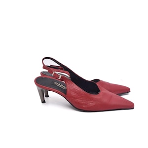 Könnte beinhalten: Ein Paar rote Slingback-Heels aus Leder mit spitzer Zehenpartie. Die Absätze haben einen silberfarbenen Absatz und einen Riemen, der sich um den Knöchel schnallt. Die Schuhe stehen auf weißem Hintergrund.