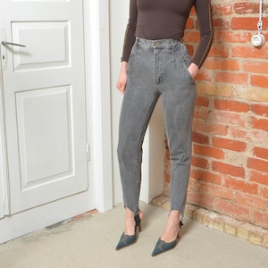 May include: Gray denim jeans with a high waist and a tapered leg. The jeans have a raw hem detail at the ankle. The model is wearing a brown long-sleeve shirt and blue heels. The background includes a white door and a brick wall.