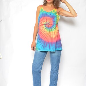 Puede incluir: Una mujer con un top sin mangas de tie-dye con un patr&oacute;n de espiral arco&iacute;ris y pantalones vaqueros azules. Lleva zapatos azules de tac&oacute;n.