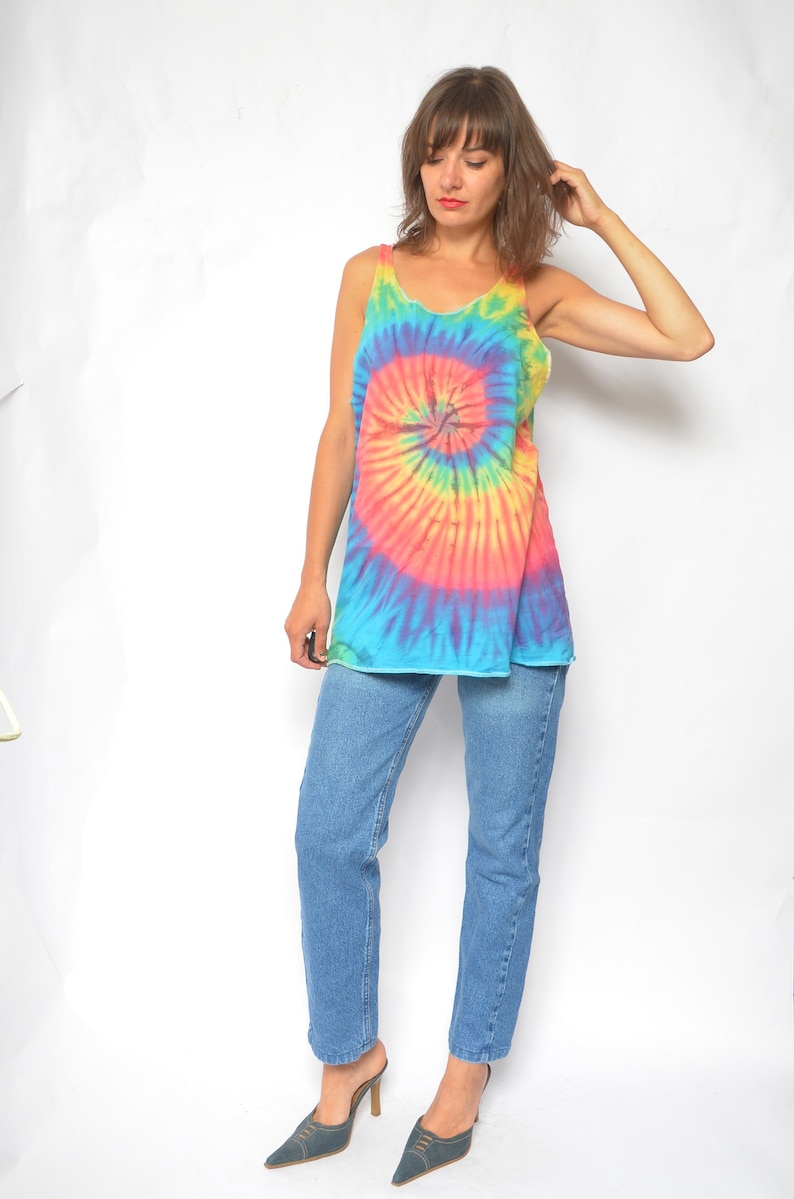 Puede incluir: Una mujer con un top sin mangas de tie-dye con un patr&oacute;n de espiral arco&iacute;ris y pantalones vaqueros azules. Lleva zapatos azules de tac&oacute;n.